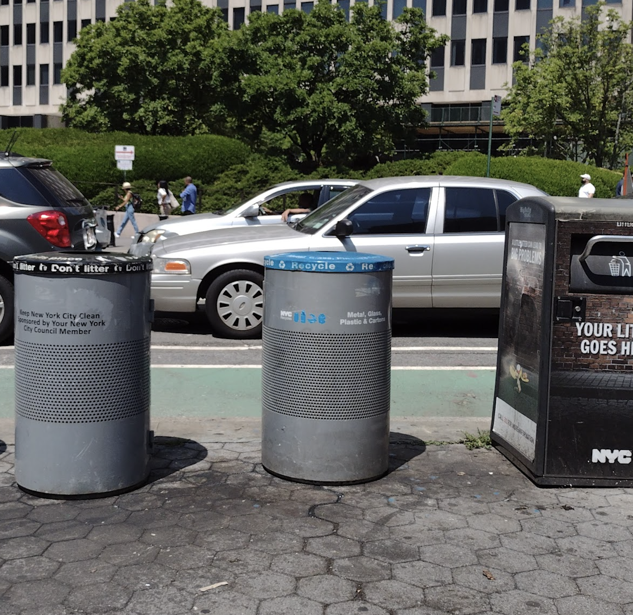 Round, grey, enclosed trash baskets are lined up next to each other. These three baskets have a small circular opening at the top and take different types of trash (mixed paper, litter, and metal/glass/plastic/cartons) A fourth basket that has a handle to open the trash basket is next to the other baskets.