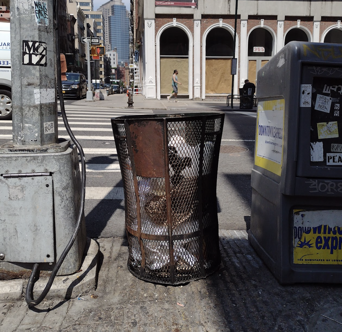 Open wire basket with some trash. Next to a pole and plastic newspaper stand 